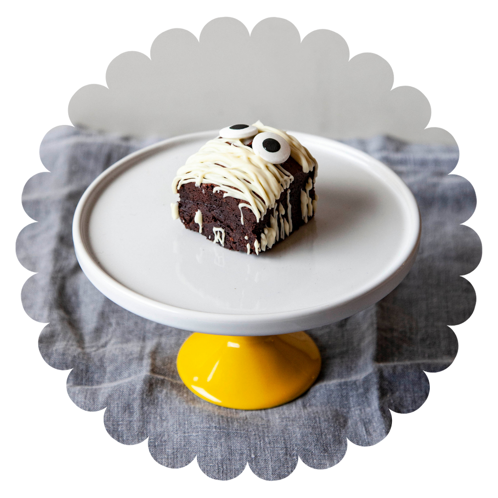 A Halloween gluten free double chocolate brownie with white chocolate drizzle and fake eyes on a round white plate, with a yellow pedestal underneath. The brownie is on a grey cloth background.