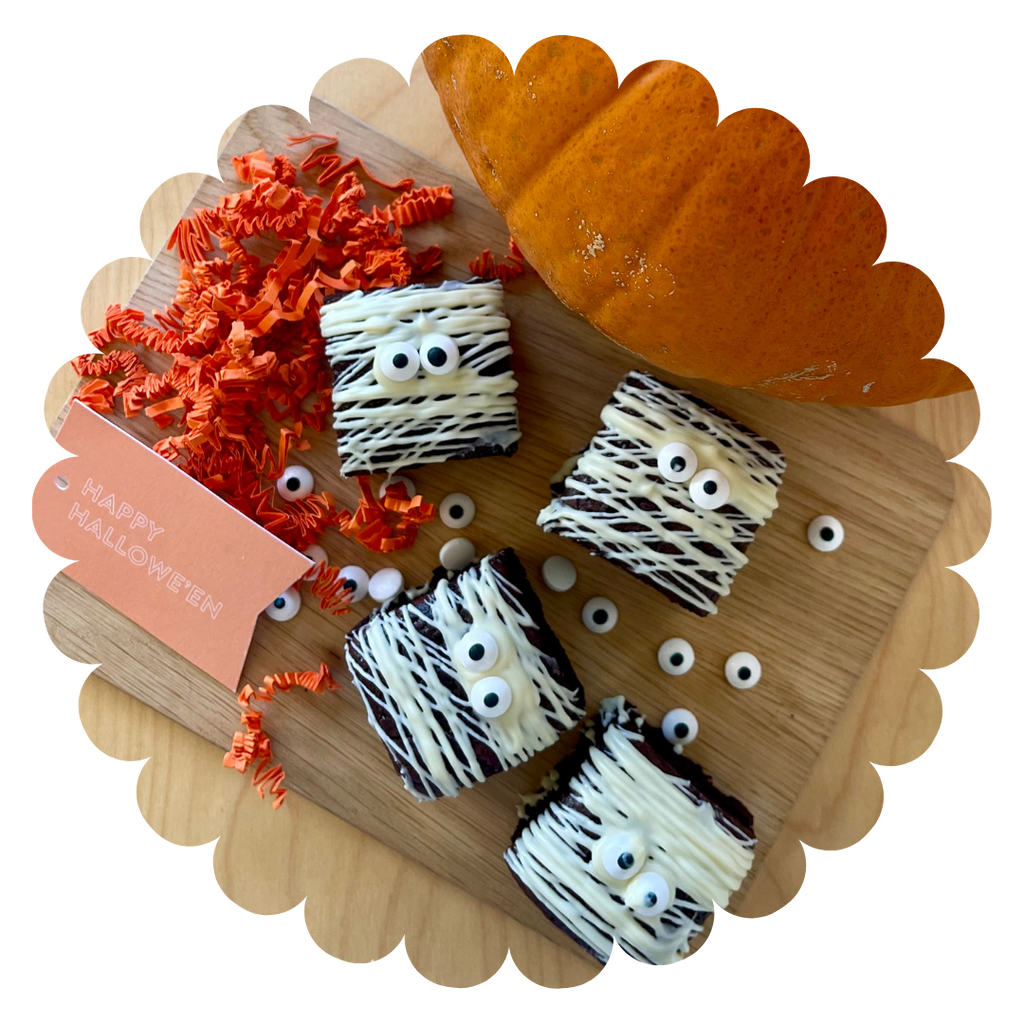 4 double chocolate Halloween gluten-free brownies with white chocolate drizzle to look like bandages and googly eyes. On a tray with a pumpkin and Happy Halloween gift tag